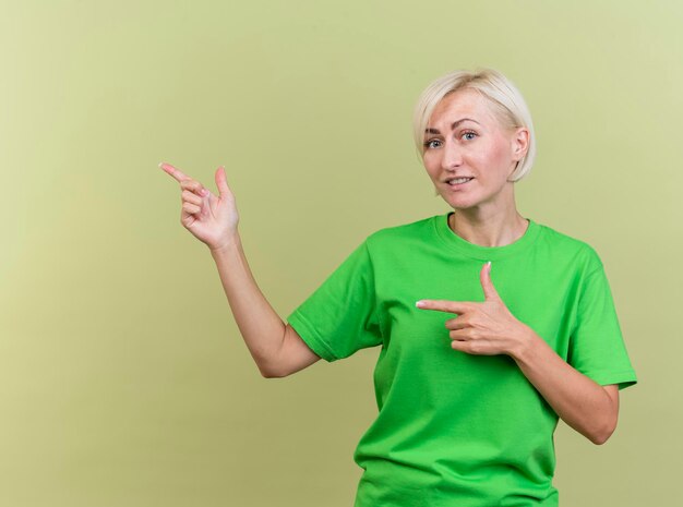 Heureux d'âge moyen blonde femme slave regardant avant pointant sur le côté isolé sur mur vert olive avec espace copie