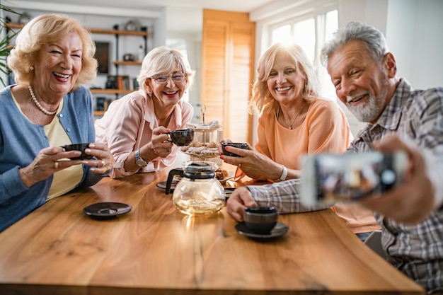 Heureux amis matures s'amusant tout en utilisant un téléphone portable et en parlant de selfie à la maison