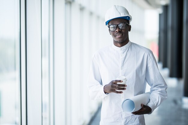 Heureux architecte afro-américain en casque tenant le projet de conception près d'un bâtiment moderne