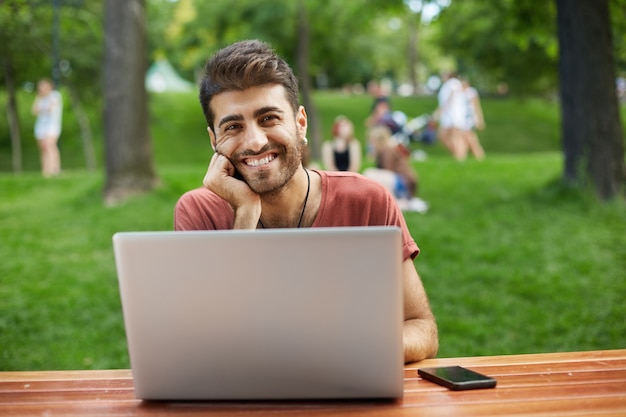 Heureux beau développeur masculin, pigiste de gars assis dans le parc avec ordinateur portable et souriant