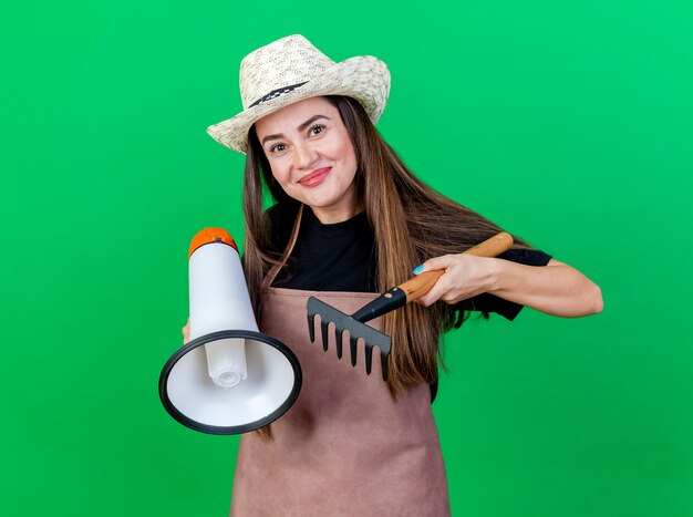 Heureux beau jardinier fille en uniforme portant chapeau de jardinage tenant haut-parleur avec râteau isolé sur vert