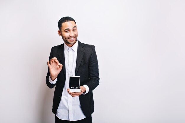 Heureux beau mec en chemise blanche de luxe, veste noire tenant un cadeau et souriant. Temps de vacances, surprise, ornementation, bonne humeur.