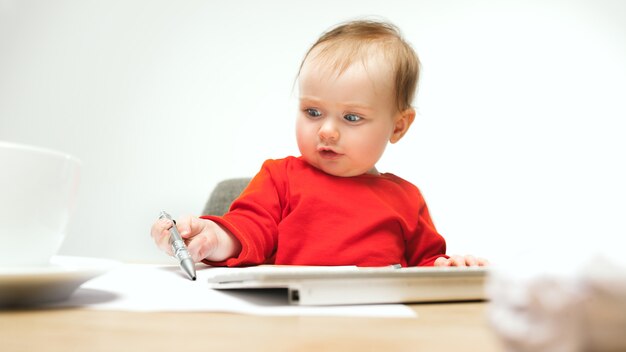 Heureux bébé fille assise avec un stylo et un clavier d'ordinateur moderne ou un ordinateur portable isolé sur un studio blanc.