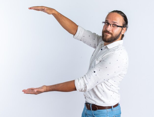 Heureux bel homme adulte portant des lunettes debout dans la vue de profil faisant un geste de taille regardant la caméra isolée sur un mur blanc