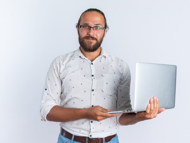 Heureux bel homme adulte portant des lunettes tenant un ordinateur portable regardant la caméra isolée sur un mur blanc