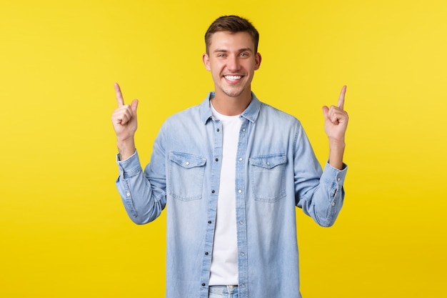 Heureux bel homme adulte avec un sourire blanc parfait, pointant les doigts vers le haut, démontrant une bannière, faisant de la publicité et recommandant d'acheter un produit, une remise spéciale d'été, un fond jaune debout.