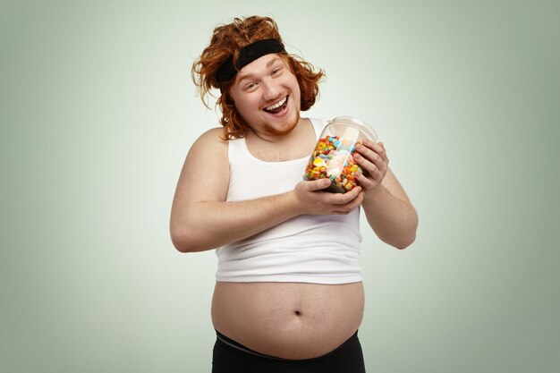 Heureux bel homme aux cheveux roux portant un bandeau et un débardeur rétréci tenant un pot en verre de bonbons, se réjouissant d'un pied de poubelle délicieux mais malsain après un entraînement cardio, luttant contre l'excès de poids