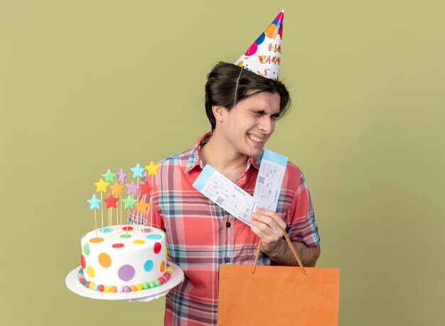 Heureux bel homme caucasien portant une casquette d'anniversaire détient des billets d'avion de sac à provisions en papier et un gâteau d'anniversaire