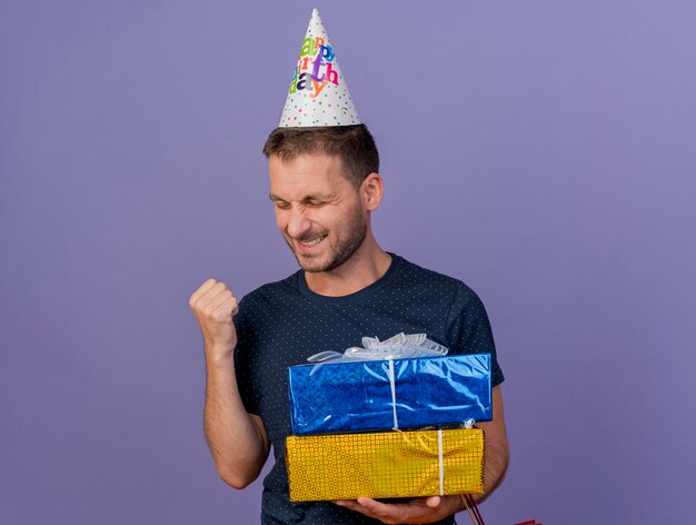 Heureux bel homme caucasien portant une casquette d'anniversaire garde le poing et détient des coffrets cadeaux isolés sur fond violet avec espace de copie