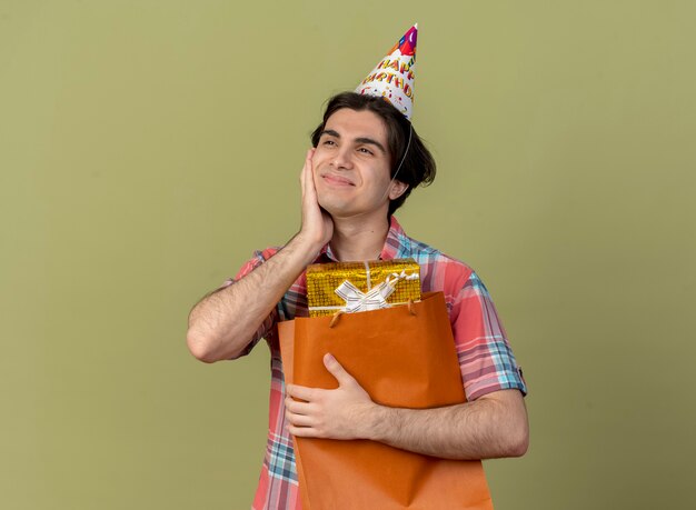 Heureux bel homme caucasien portant une casquette d'anniversaire met la main sur le visage et tient une boîte-cadeau dans un sac à provisions en papier