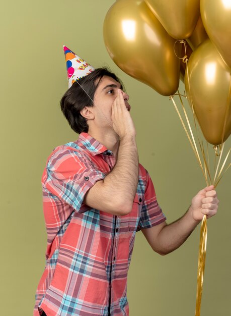 Heureux bel homme caucasien portant une casquette d'anniversaire se tient sur le côté tenant la main près de la bouche en regardant des ballons à l'hélium