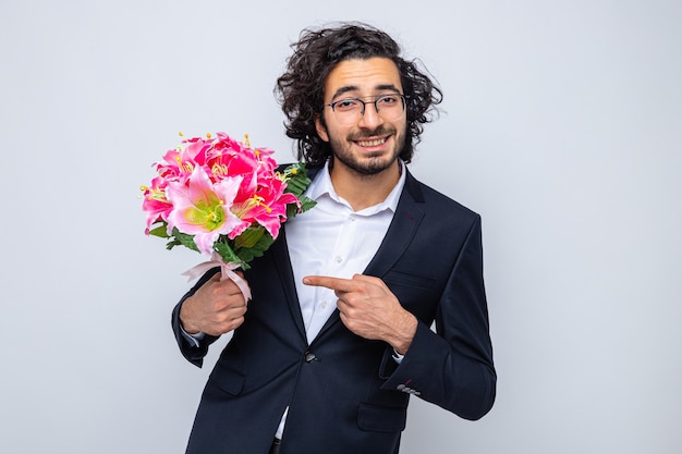 Heureux bel homme en costume avec bouquet de fleurs pointant avec l'index en regardant la caméra souriant joyeusement célébrant la journée internationale de la femme le 8 mars debout sur fond blanc