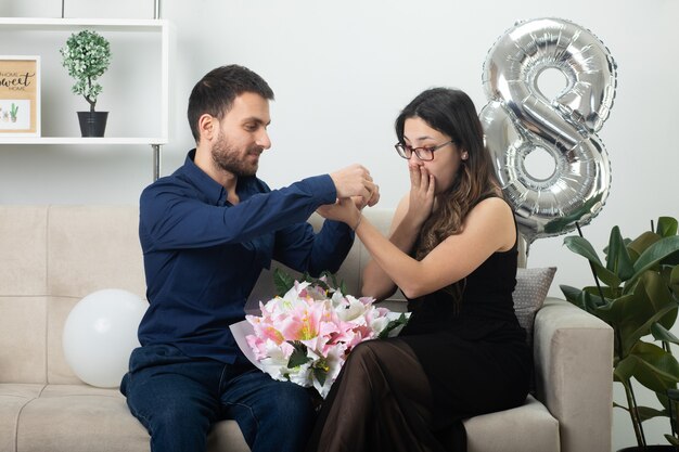 Heureux bel homme mettant le bracelet à portée de main d'une jolie jeune femme excitée dans des lunettes optiques tenant un bouquet de fleurs assis sur un canapé dans le salon le jour de la journée internationale de la femme en mars