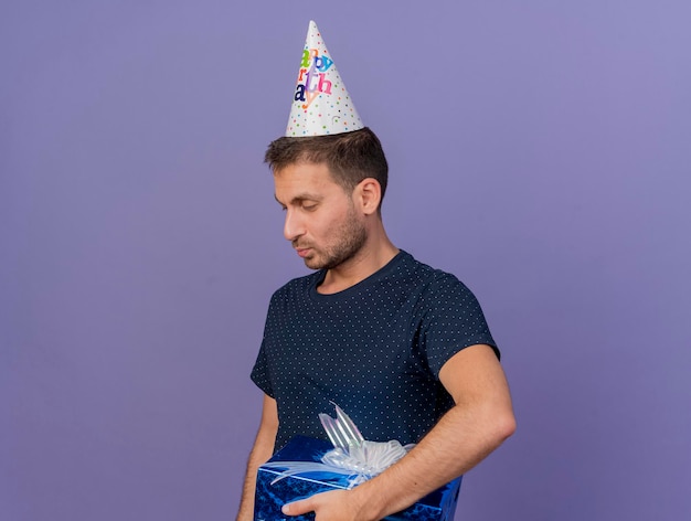 Heureux bel homme portant une casquette d'anniversaire détient une boîte-cadeau à côté isolé sur mur violet avec espace copie