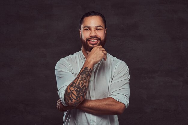 Heureux bel homme tatoué afro-américain avec des cheveux et une barbe élégants dans une chemise blanche, tenant la main sur son menton.