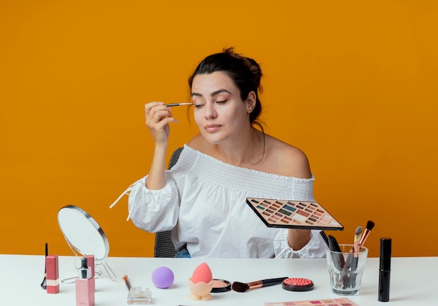 Heureux belle fille est assise à table avec des outils de maquillage se penche sur l'application de miroir de fard à paupières avec pinceau de maquillage isolé sur mur orange