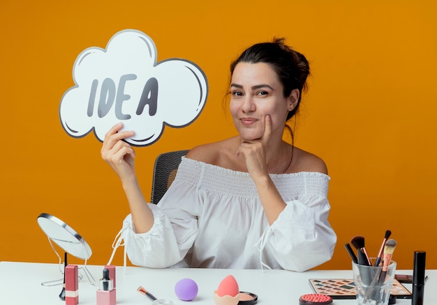 Heureux belle fille est assise à table avec des outils de maquillage tenant la marque idée à isolé sur mur orange