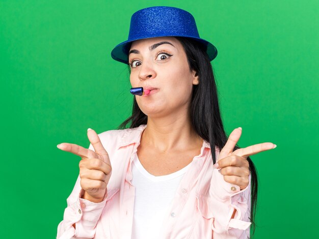 Heureux belle jeune femme portant un chapeau de fête soufflant des points de sifflet de fête à l'avant isolés sur un mur vert