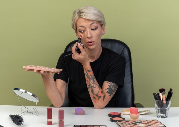 Heureux belle jeune fille assise à table avec des outils de maquillage appliquant un fard à paupières avec un pinceau de maquillage isolé sur un mur vert olive