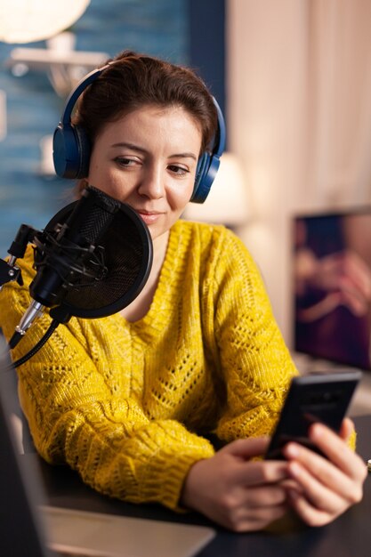 Heureux blogueur lisant des messages de fans utilisant un smartphone assis dans un studio de podcast à domicile pendant la diffusion en direct, l'enregistrement