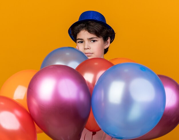 Heureux à la caméra petit garçon portant un chapeau de fête bleu debout derrière des ballons