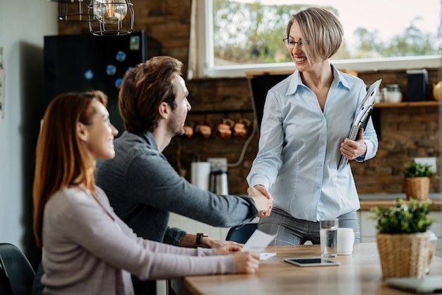 Heureux conseiller financier saluant un jeune couple à la maison et serrant la main d'un homme.