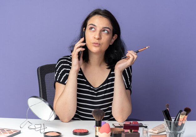 Heureux côté jeune belle fille assise à table avec des outils de maquillage parle au téléphone tenant un pinceau de maquillage isolé sur un mur bleu