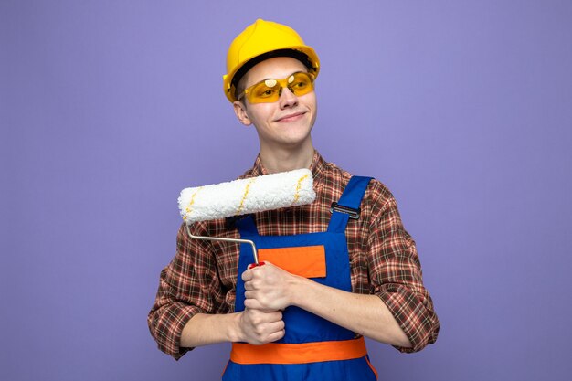 Heureux côté jeune constructeur masculin portant des uniformes et des lunettes tenant une brosse à rouleau