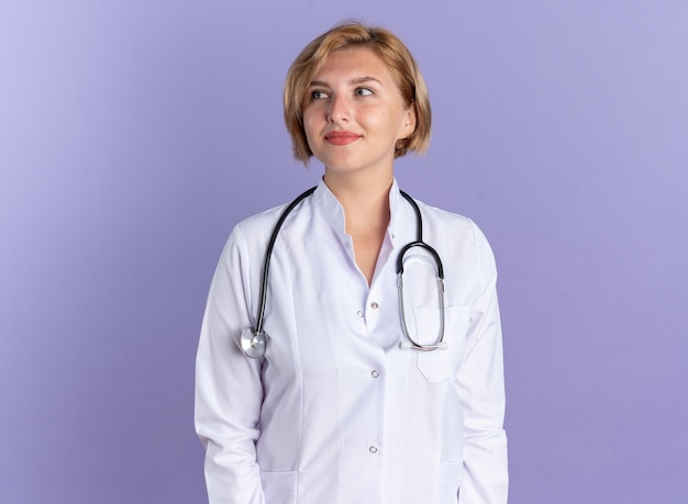 Heureux côté jeune femme médecin portant une robe médicale avec stéthoscope isolé sur mur bleu