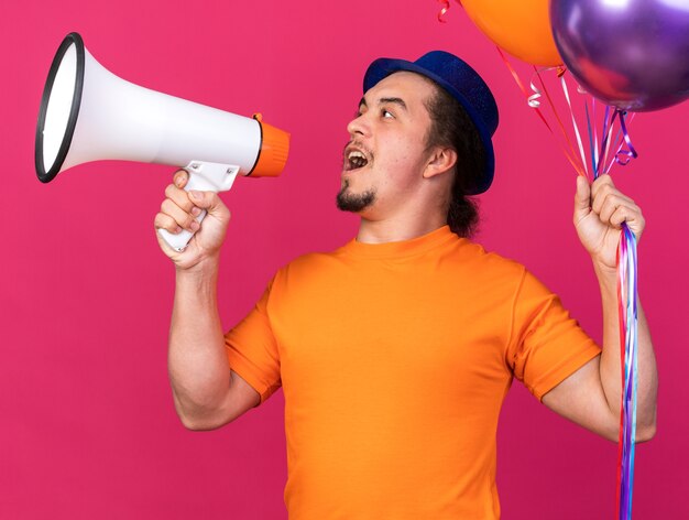 Heureux à côté d'un jeune homme portant un chapeau de fête tenant des ballons parle sur un haut-parleur isolé sur un mur rose
