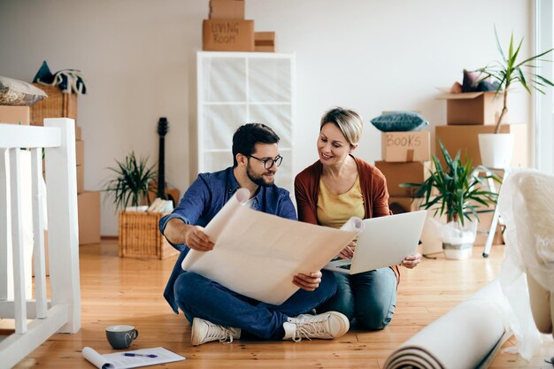 Heureux couple examinant des plans et utilisant un ordinateur portable dans leur nouvel appartement