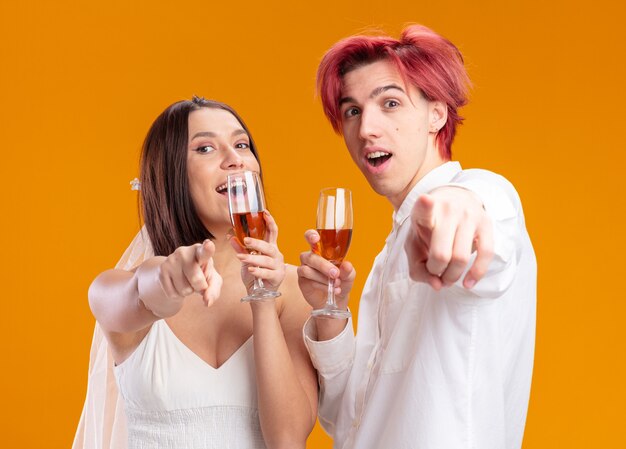 Heureux couple de mariage marié et mariée en robe de mariée souriant joyeusement posant ensemble tenant des verres de champagne pointant avec l'index à l'avant