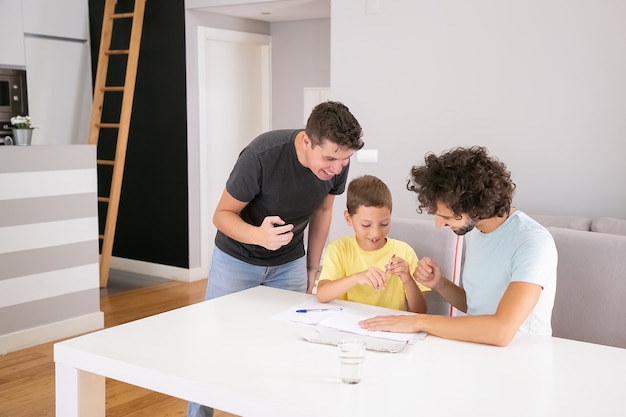 Heureux couple de papas aidant un garçon concentré avec la tâche à la maison de l'école, assis à table ensemble, écrivant sur papier. Concept de famille et de parents gays