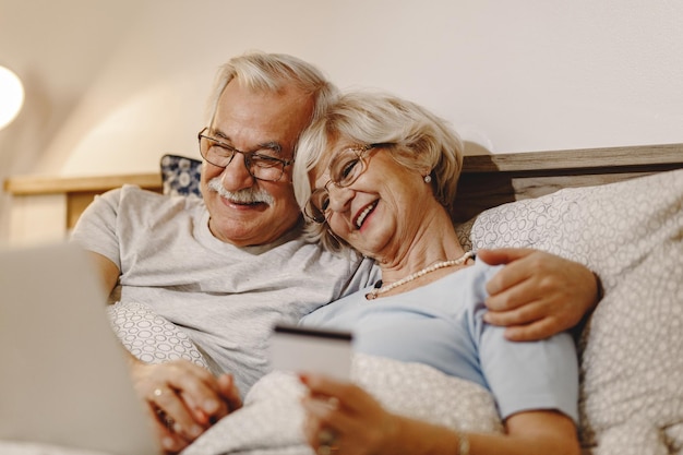 Heureux couple de personnes âgées utilisant un ordinateur portable et une carte de crédit tout en se relaxant dans la chambre