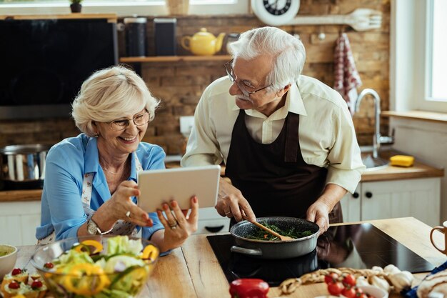 Heureux couple de personnes âgées utilisant une tablette numérique et s'amusant tout en préparant le déjeuner dans la cuisine
