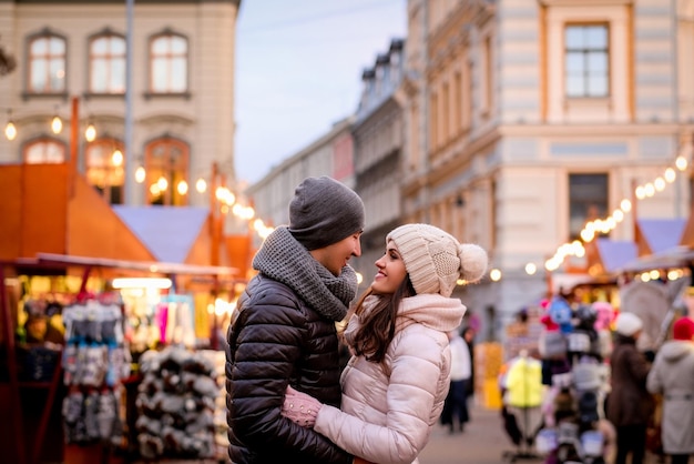 Heureux couple romantique portant des vêtements d'hiver étreignant debout dans la rue du soir avec la foire de Noël