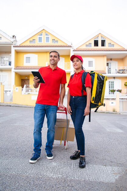 Heureux courriers debout et regardant l'adresse via tablette. Joyeux postiers livrant la commande ensemble dans un sac à dos thermo et des boîtes en carton sur chariot. Service de livraison et concept d'achat en ligne