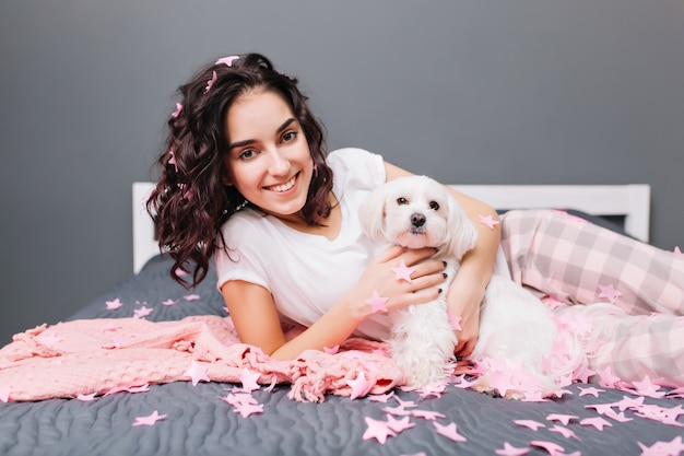 Heureux doux moments de belle jeune femme en pyjama avec des cheveux bouclés brune coupée se détendre sur le lit avec petit chien dans un appartement moderne. Sourire en guirlandes roses, se détendre à la maison confort