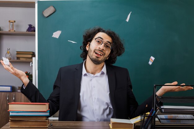 Heureux écartant les mains enseignant portant des lunettes larmes papier assis à table avec des outils scolaires en classe