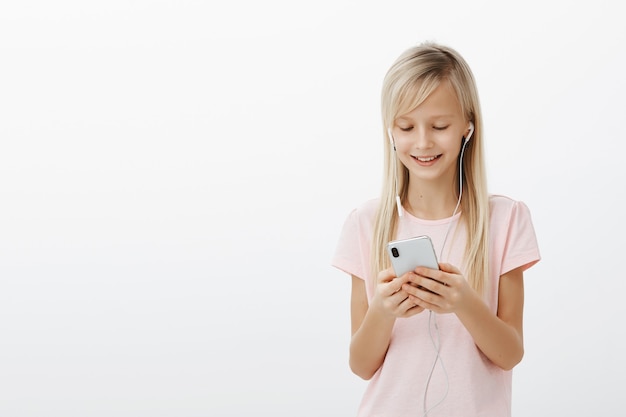 Heureux enfant créatif regardant des vidéos en attendant maman. Portrait d'adorable jeune fille aux cheveux blonds, écoutant de la musique dans des écouteurs et tenant un smartphone, souriant d'un message positif