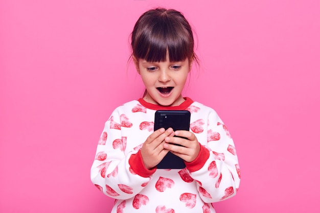 Heureux enfant de sexe féminin excité portant un cavalier avec des coeurs tenant le téléphone portable dans les mains et voit quelque chose d'étonnant à son écran, garde la bouche ouverte, posant isolé sur un mur rose.