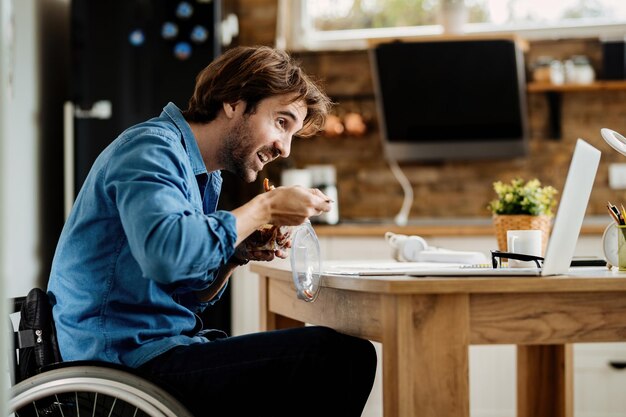 Heureux entrepreneur en fauteuil roulant mangeant en lisant un e-mail sur un ordinateur portable à la maison.