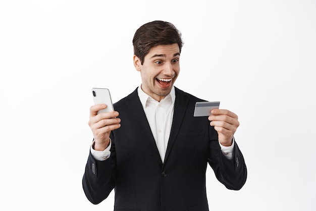 Heureux entrepreneur regarde la carte de crédit avec un visage étonné payant en ligne avec un smartphone debout en costume noir sur fond blanc