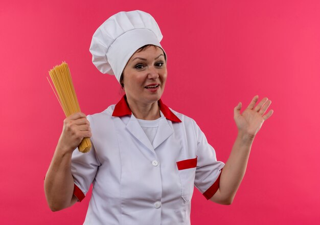 Heureux femme d'âge moyen cuisinier en uniforme de chef holding spaghetti points sur le côté sur un mur rose isolé