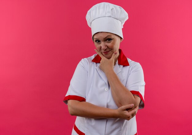 Heureux femme d'âge moyen cuisinier en uniforme de chef a mis sa main sur le menton sur un mur rose isolé avec copie espace