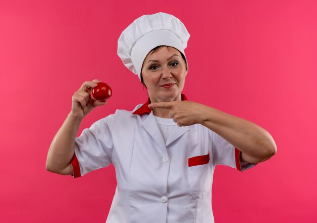 Heureux femme d'âge moyen cuisinier en uniforme de chef pointe le doigt à la tomate dans sa main sur un mur rose isolé