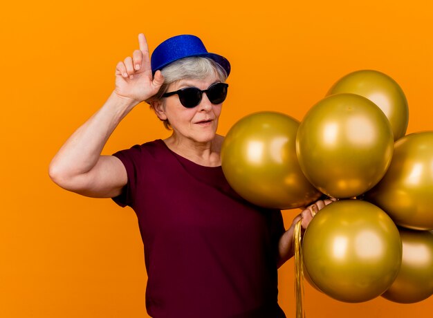 Heureux femme âgée à lunettes de soleil portant chapeau de fête se dresse avec des ballons d'hélium pointant vers le haut isolé sur un mur orange avec copie espace