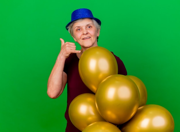 Heureux femme âgée portant chapeau de fête se dresse avec des ballons d'hélium faisant des gestes appelez-moi signe sur vert