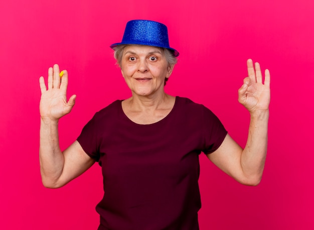 Heureux femme âgée portant chapeau de fête se dresse avec la main levée tenant le sifflet et gesticulant signe de la main ok isolé sur mur rose