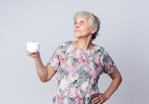 Heureux femme âgée tient la tasse et met la main sur la taille en levant isolé sur mur blanc